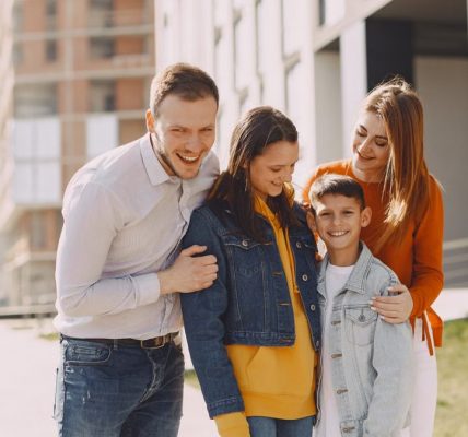 Delighted family in trendy casual wear holding each other and standing near modern building on city street while walking together during weekend